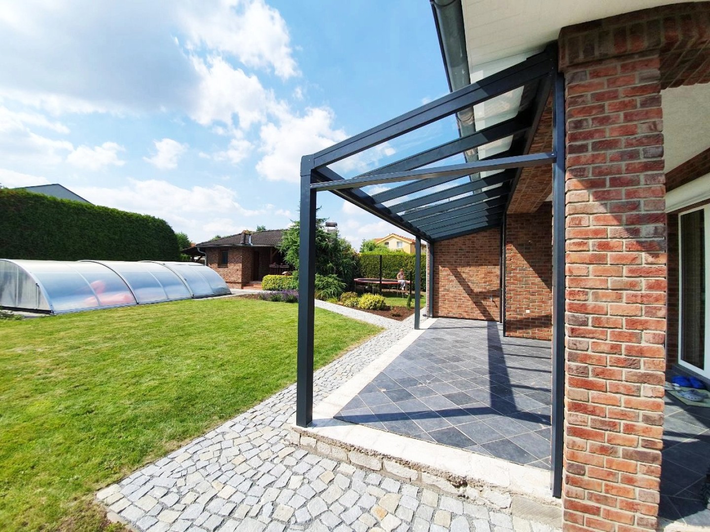Une pergola moderne en aluminium anthracite avec un toit en verre, près d'une maison en briques dotée d'un jardin et d'une piscine.