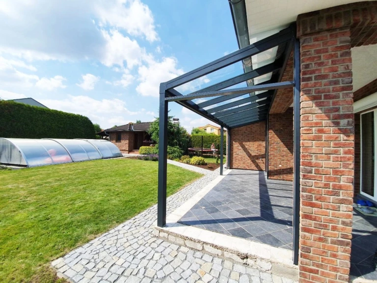 Une pergola moderne en aluminium anthracite avec un toit en verre, près d'une maison en briques dotée d'un jardin et d'une piscine.
