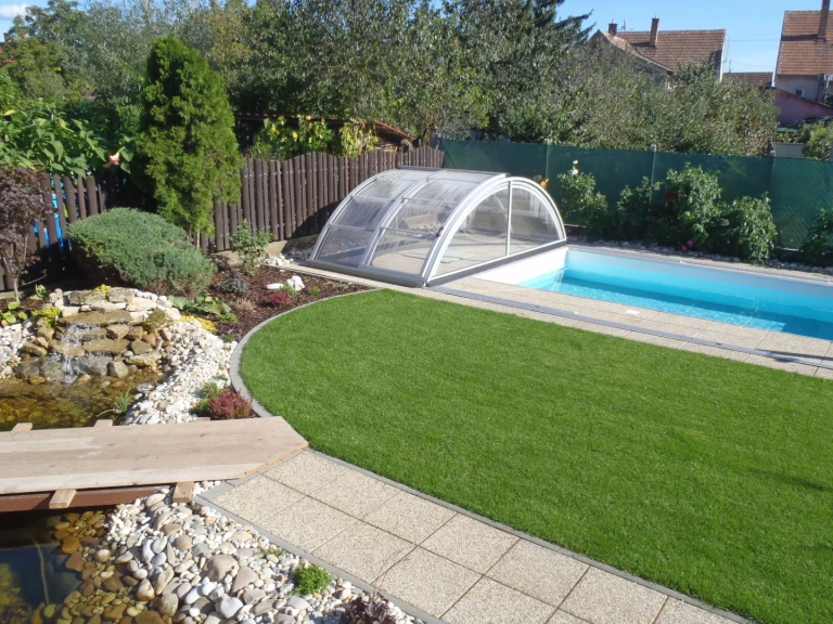Une piscine KLASIK moderne à toit bas, avec une eau bleu clair et un jardin aménagé comprenant un petit étang.
