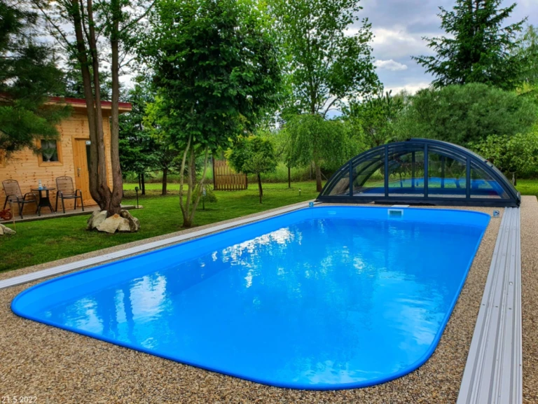 Une piscine encastrée bleue dans le jardin, avec un abri partiellement ouvert et une maisonnette en bois en arrière-plan