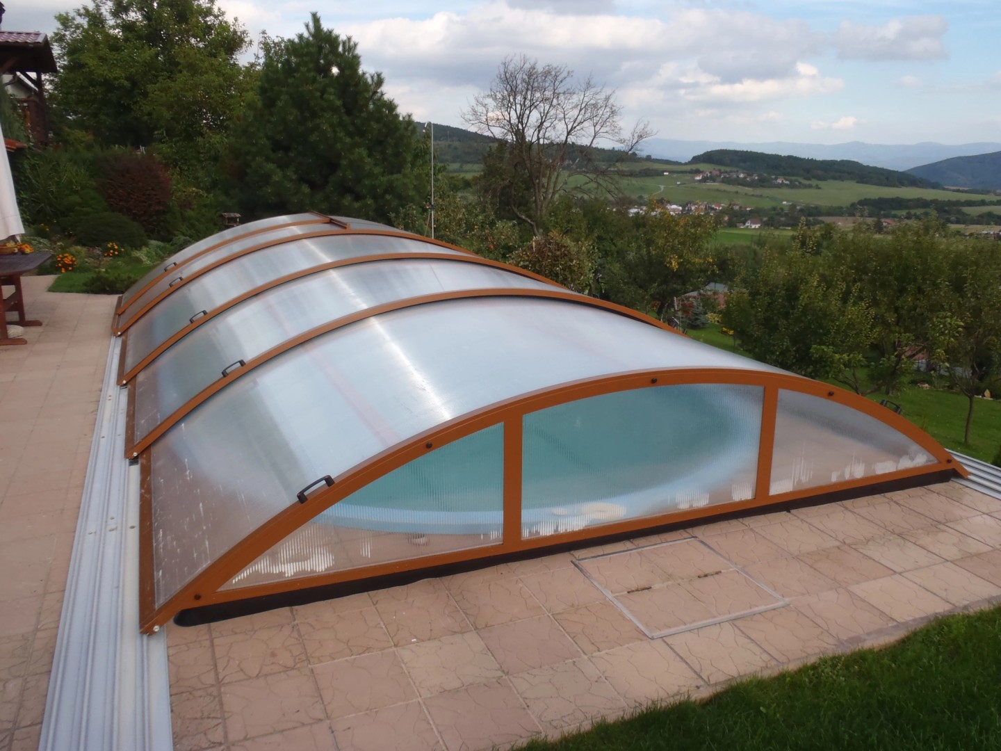Une petite verrière arquée fermée, dotée d'un cadre marron, surplombant la piscine et située sur une terrasse offrant une vue sur un paysage vallonné.