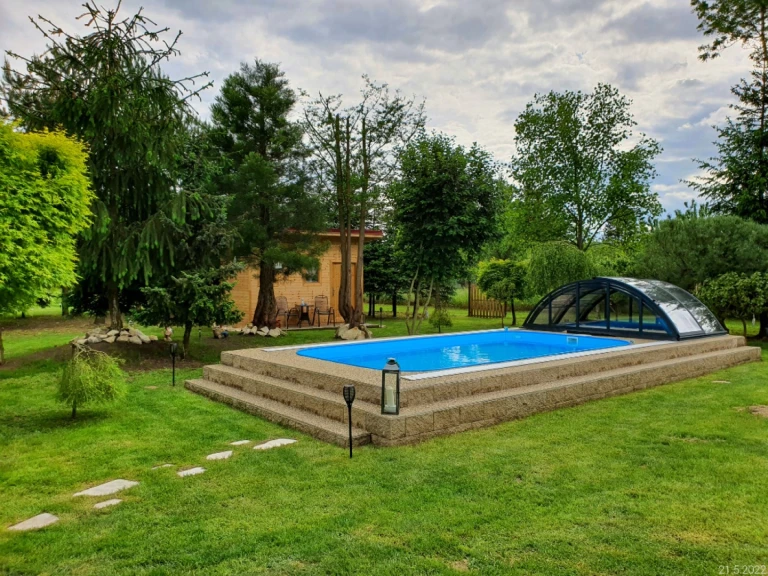 Une piscine de jardin avec un liner bleu et une couverture coulissante basse, située sur une terrasse surélevée entourée de verdure.