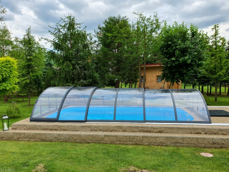 Une couverture de piscine basse et fermée, en forme d'arc, au-dessus d'une piscine bleue dans un jardin arboré.