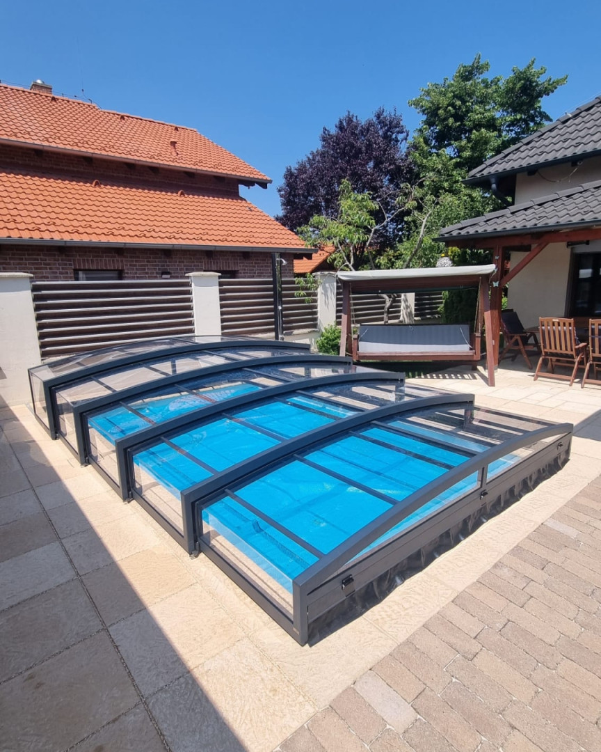 Une couverture télescopique basse de couleur anthracite pour une piscine installée sur un sol pavé dans le jardin d'une maison individuelle.