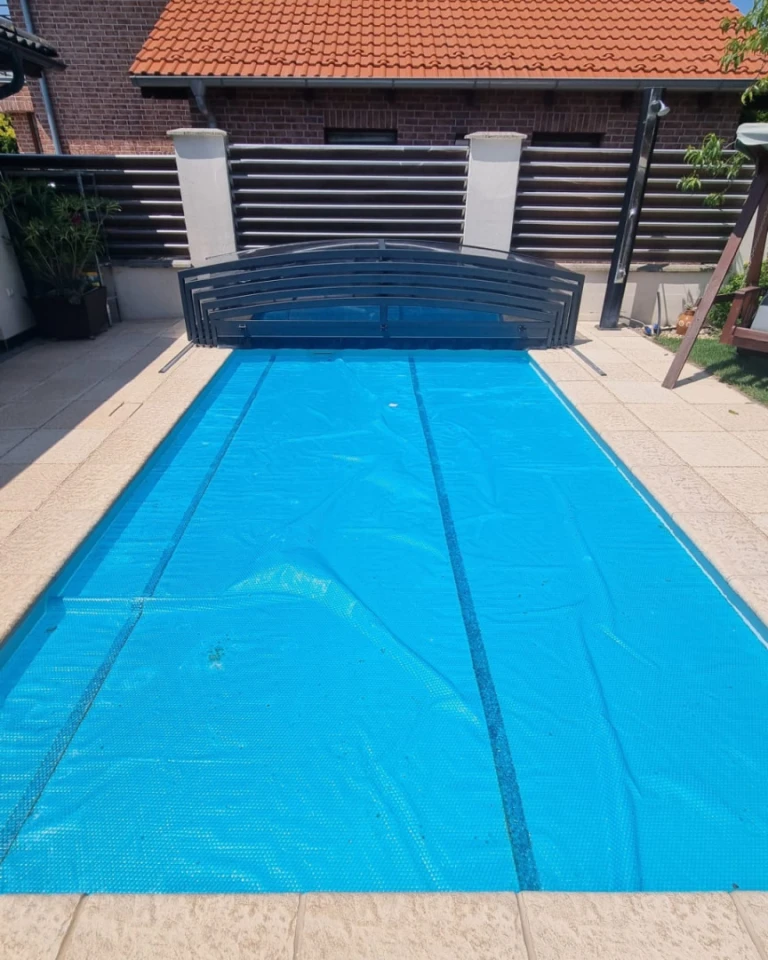 Piscine recouverte d’une bâche solaire bleue, avec une toiture coulissante ouverte sur le pourtour