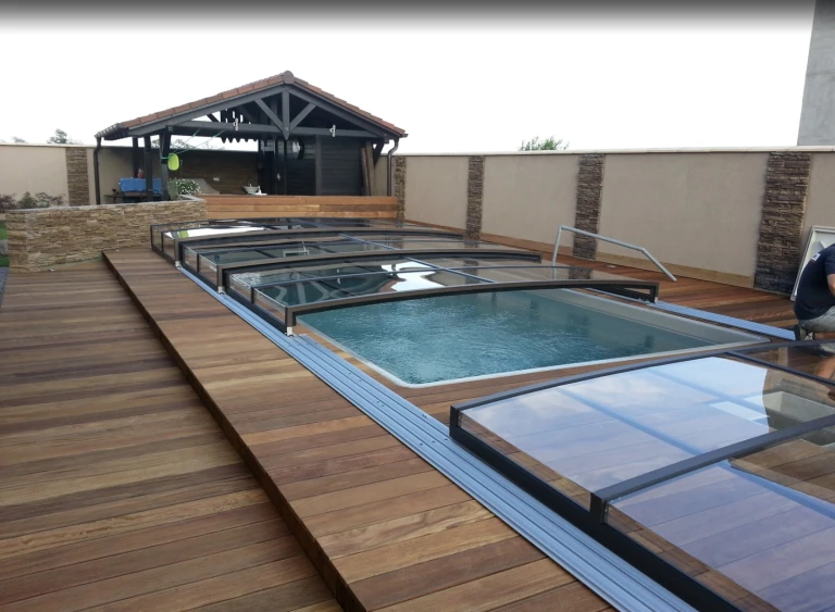 Une couverture de piscine moderne et basse, dotée de panneaux coulissants en verre, sur la terrasse en bois d'une maison individuelle.