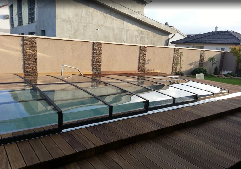 Une couverture de piscine moderne et basse, avec un cadre noir et un panneau transparent, installée sur une terrasse revêtue de bois.