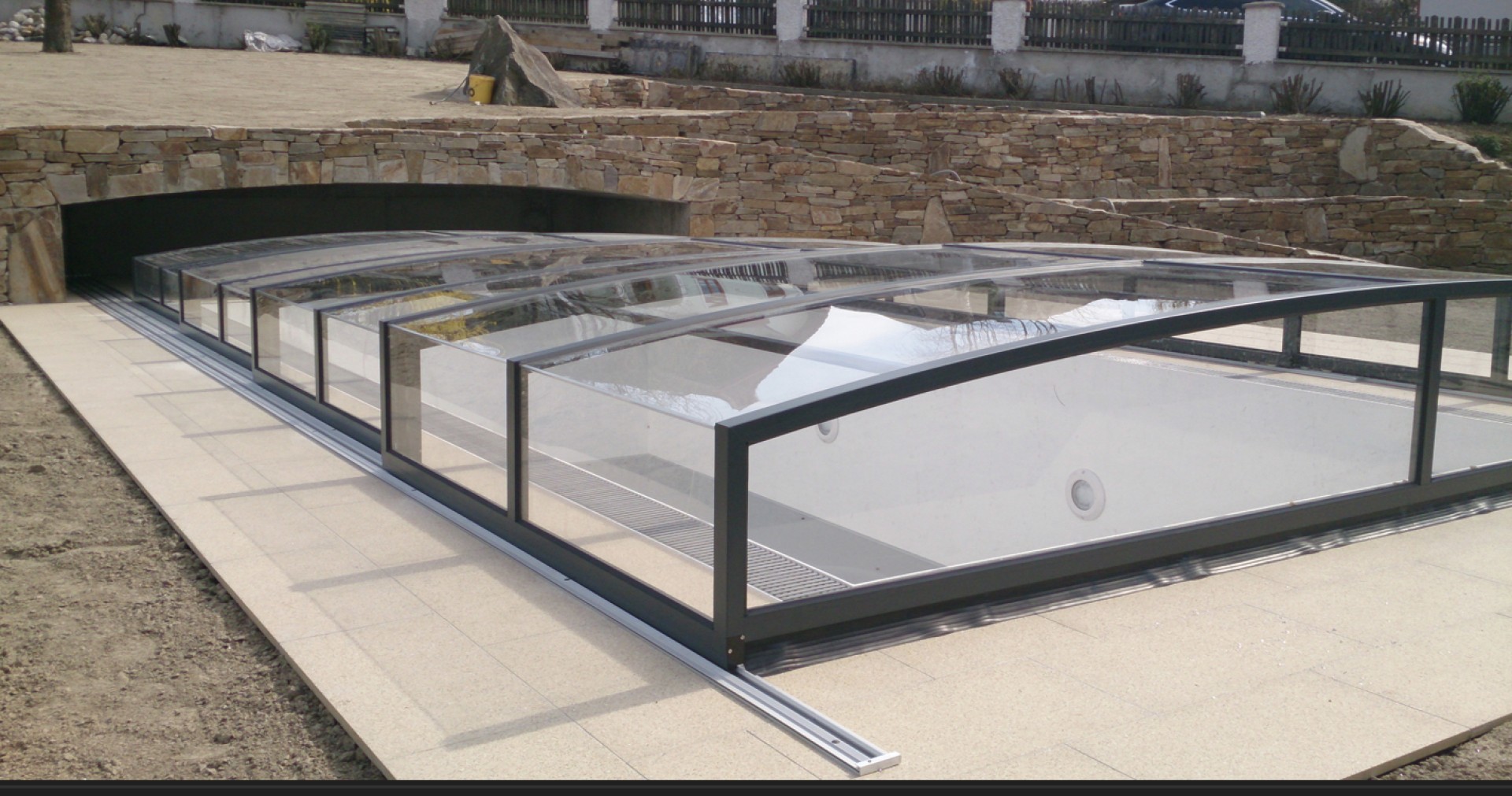 Une couverture de piscine moderne et basse, dotée d'un revêtement transparent, dans un jardin dallé de pierre.