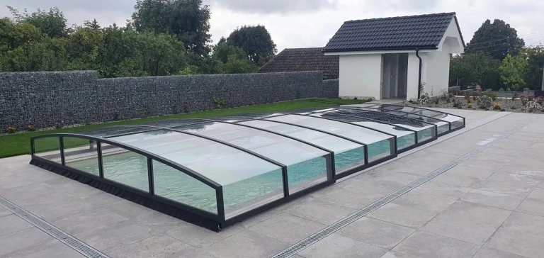 Une couverture de piscine moderne et basse, avec une structure noire et du verre transparent, installée sur la terrasse près de la maison.