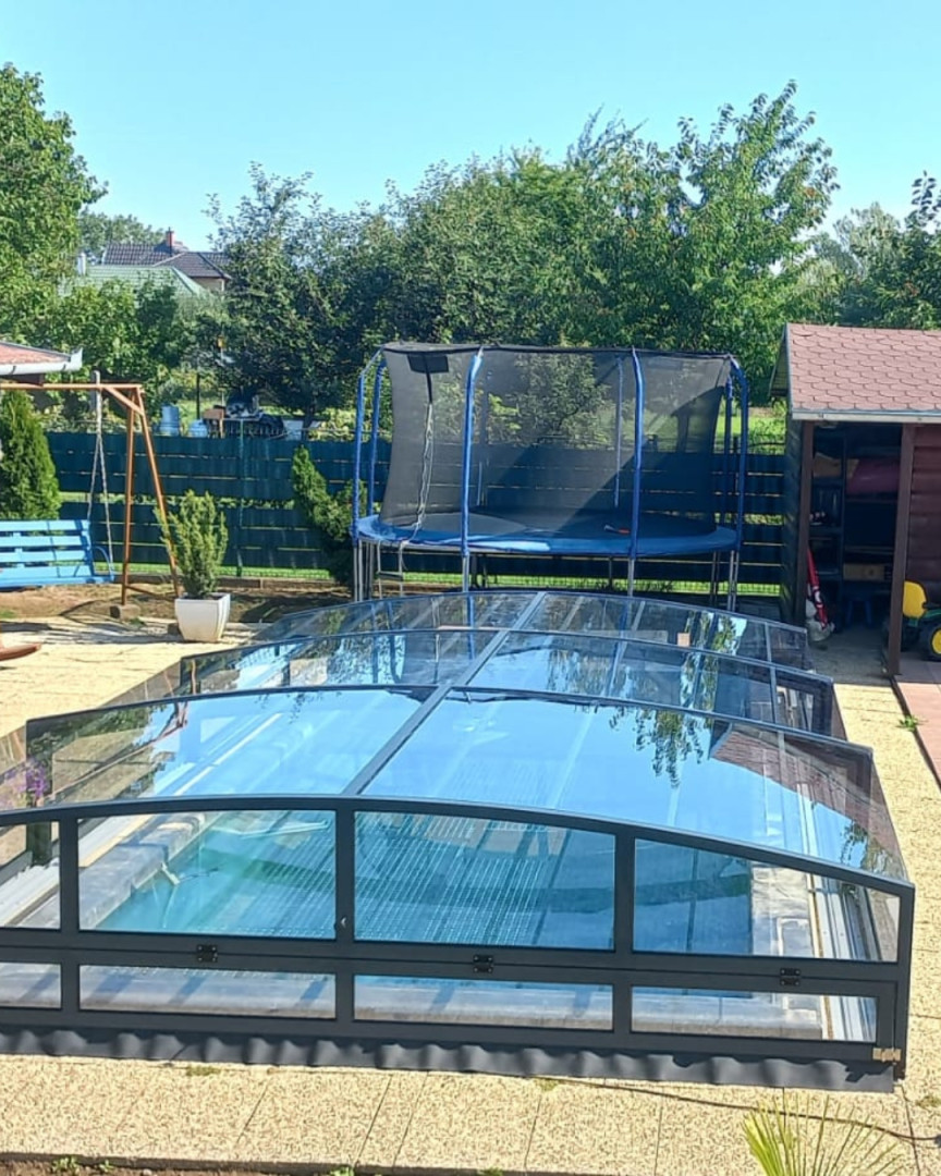 Un abri de piscine moderne et bas, avec un cadre anthracite, dans un jardin équipé d’un trampoline