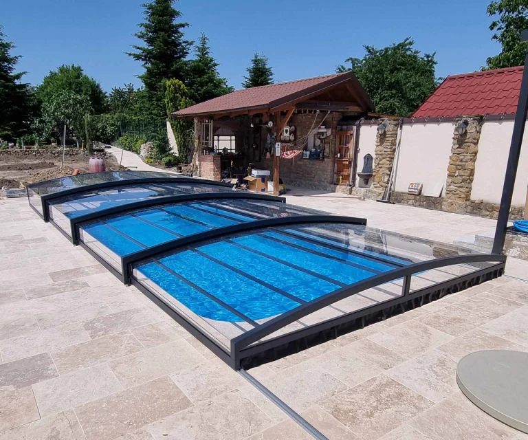 Une couverture de piscine moderne et basse, avec un cadre foncé et un panneau transparent, installée sur une terrasse en pierre.