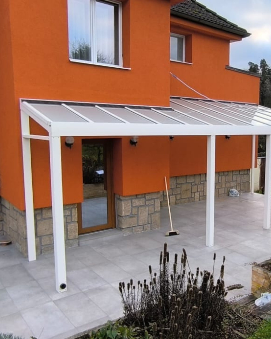 Une pergola moderne en aluminium blanc fixée à une maison orange dotée d'un toit en verre et d'un dallage