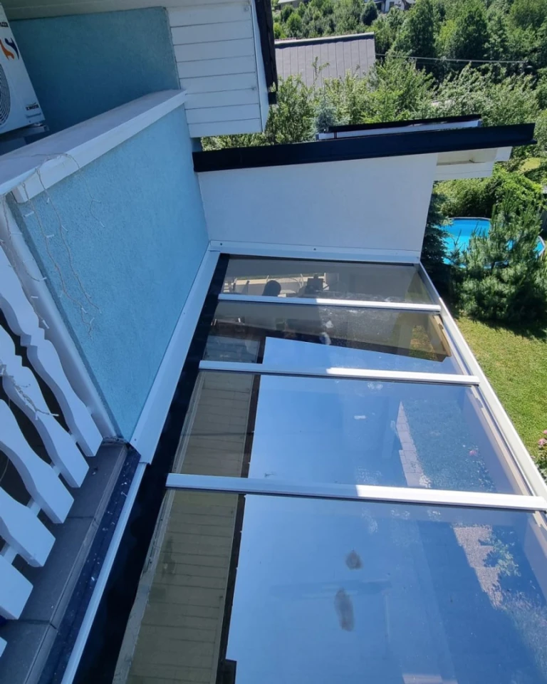 Vue d'en haut sur le toit en verre d'une pergola en aluminium fixée à la façade d'une maison, avec un jardin en arrière-plan