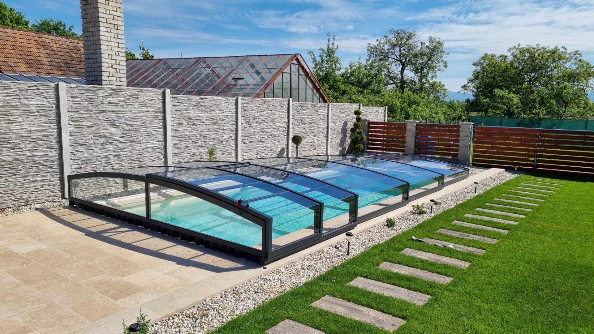 Un abri de piscine moderne et bas, avec une structure anthracite et du verre transparent, dans un jardin bien entretenu.