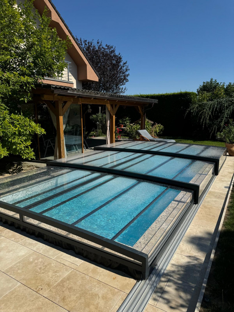 Une couverture de piscine moderne et basse, dotée d'une structure anthracite et d'un revêtement transparent, installée dans le jardin d'une maison individuelle.