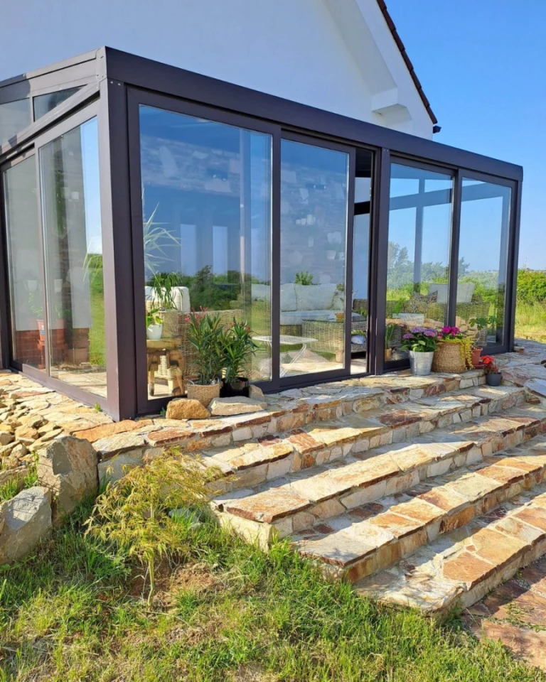 Une véranda moderne vitrée, dotée d'un cadre en aluminium foncé, construite sur une fondation en pierre avec un escalier.