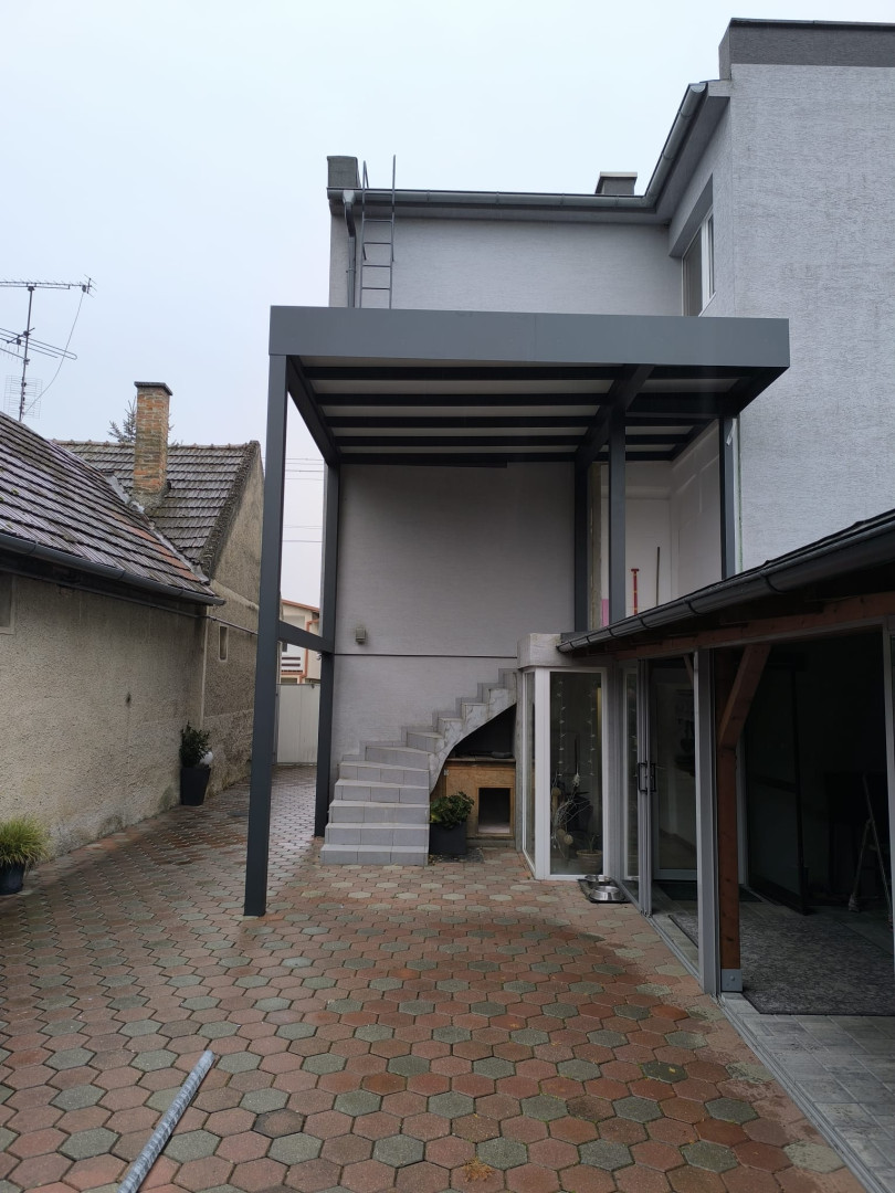 Une pergola moderne en aluminium gris, fabriquée sur mesure, installée sur la terrasse d'une maison individuelle au-dessus d'un escalier extérieur.