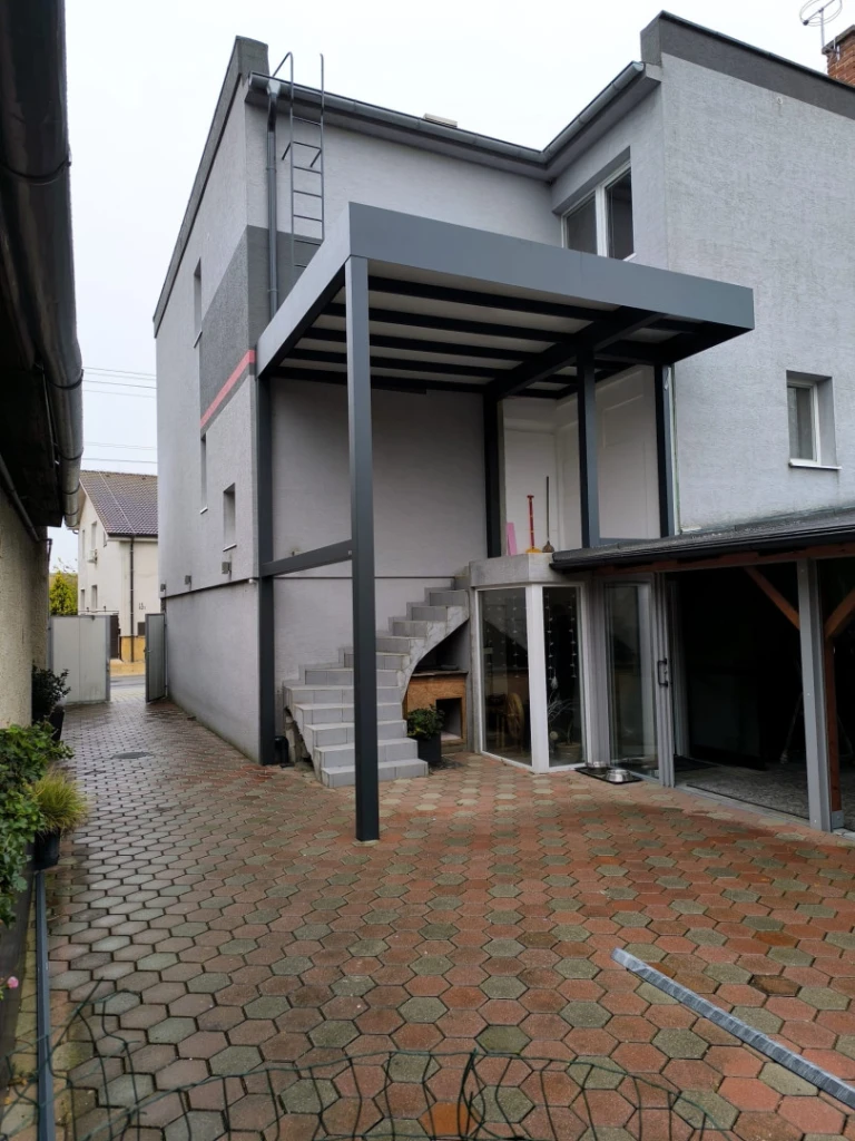 Une pergola moderne en aluminium gris foncé, dotée d'un toit fixe, fixée à la façade d'une maison individuelle au-dessus de l'escalier extérieur.