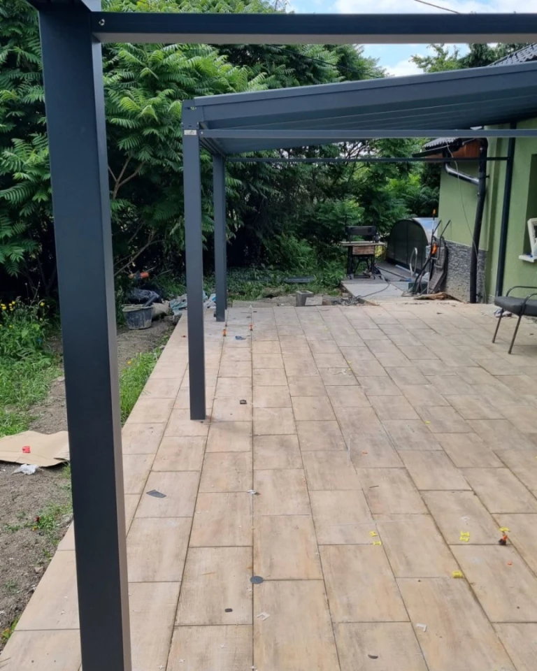Une pergola moderne en aluminium gris foncé, installée sur la terrasse pavée d'une maison individuelle donnant sur le jardin.