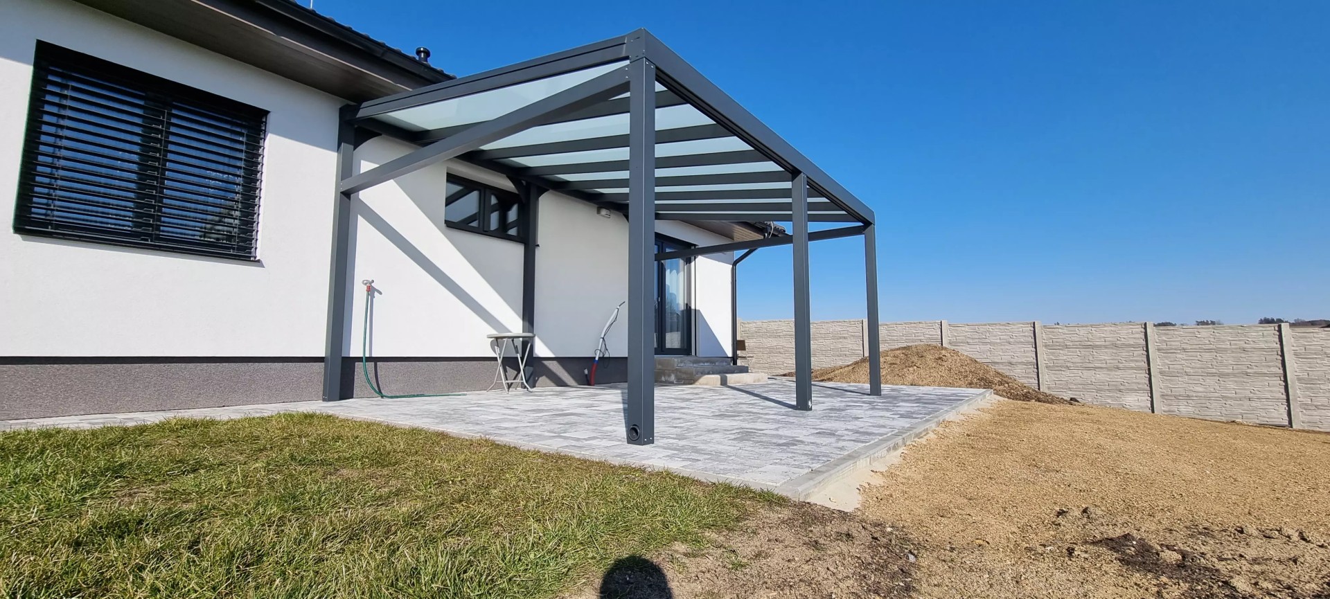 Une pergola moderne en aluminium anthracite, dotée d'un toit en verre, adossée à une maison individuelle sur une terrasse carrelée.