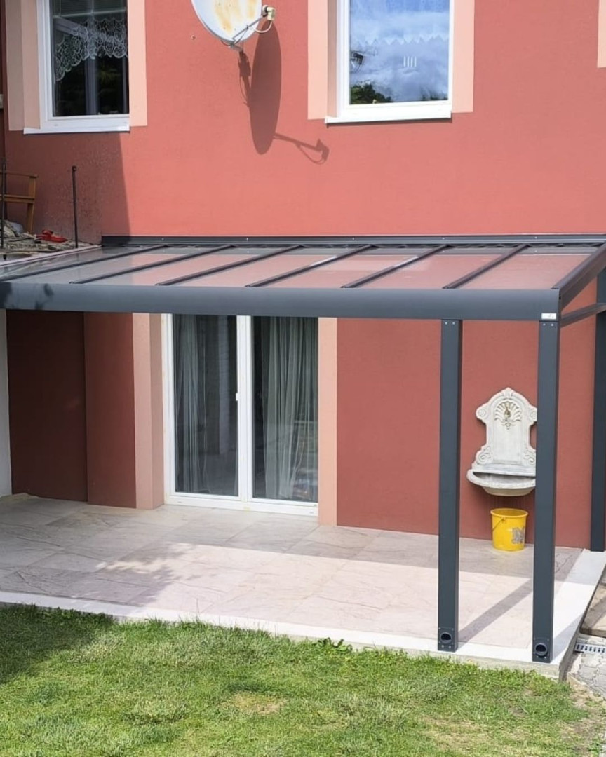 Une pergola moderne en aluminium avec un toit vitré, fixée au mur de la maison rouge au-dessus de la terrasse.