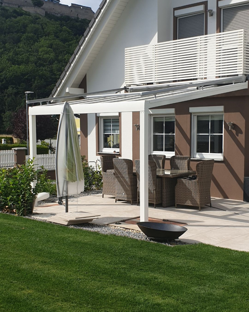 Une pergola en aluminium blanche avec un toit vitré sur la terrasse d'une maison individuelle, équipée d'un coin salon et offrant une vue sur le château en arrière-plan.