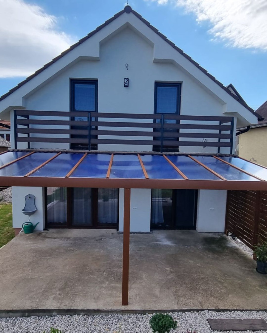 Une pergola en aluminium marron avec un toit en verre, adossée à une maison individuelle dotée d'un balcon.