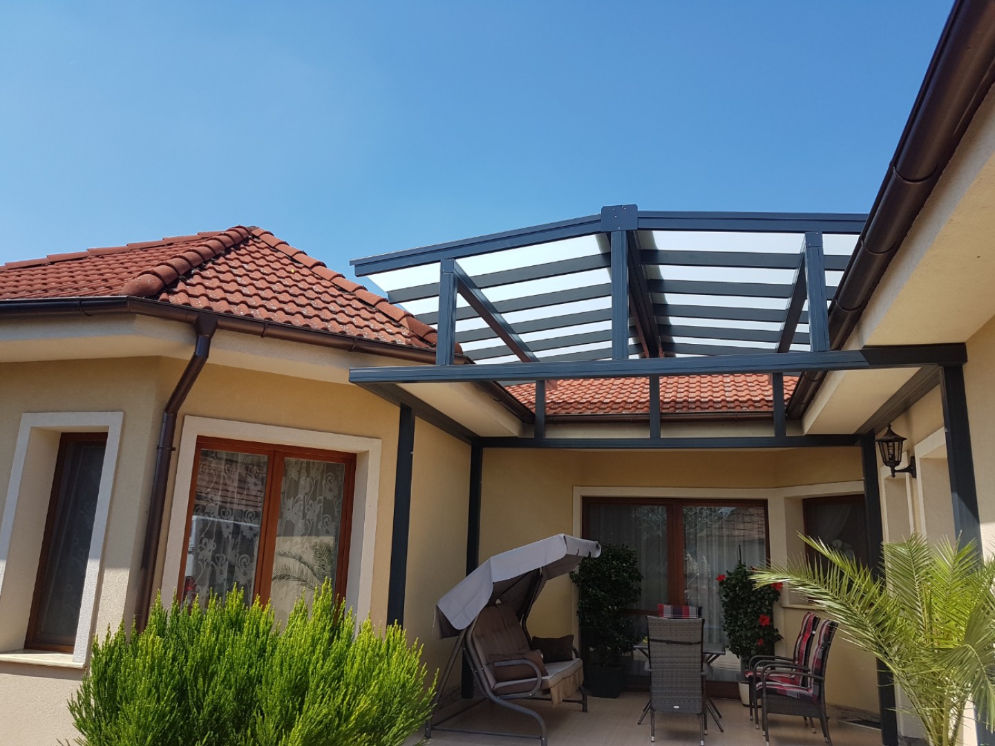 Pergola moderne en aluminium ROBUST avec toit vitré, attenante à une maison individuelle, équipée de mobilier de terrasse et d'une balançoire.