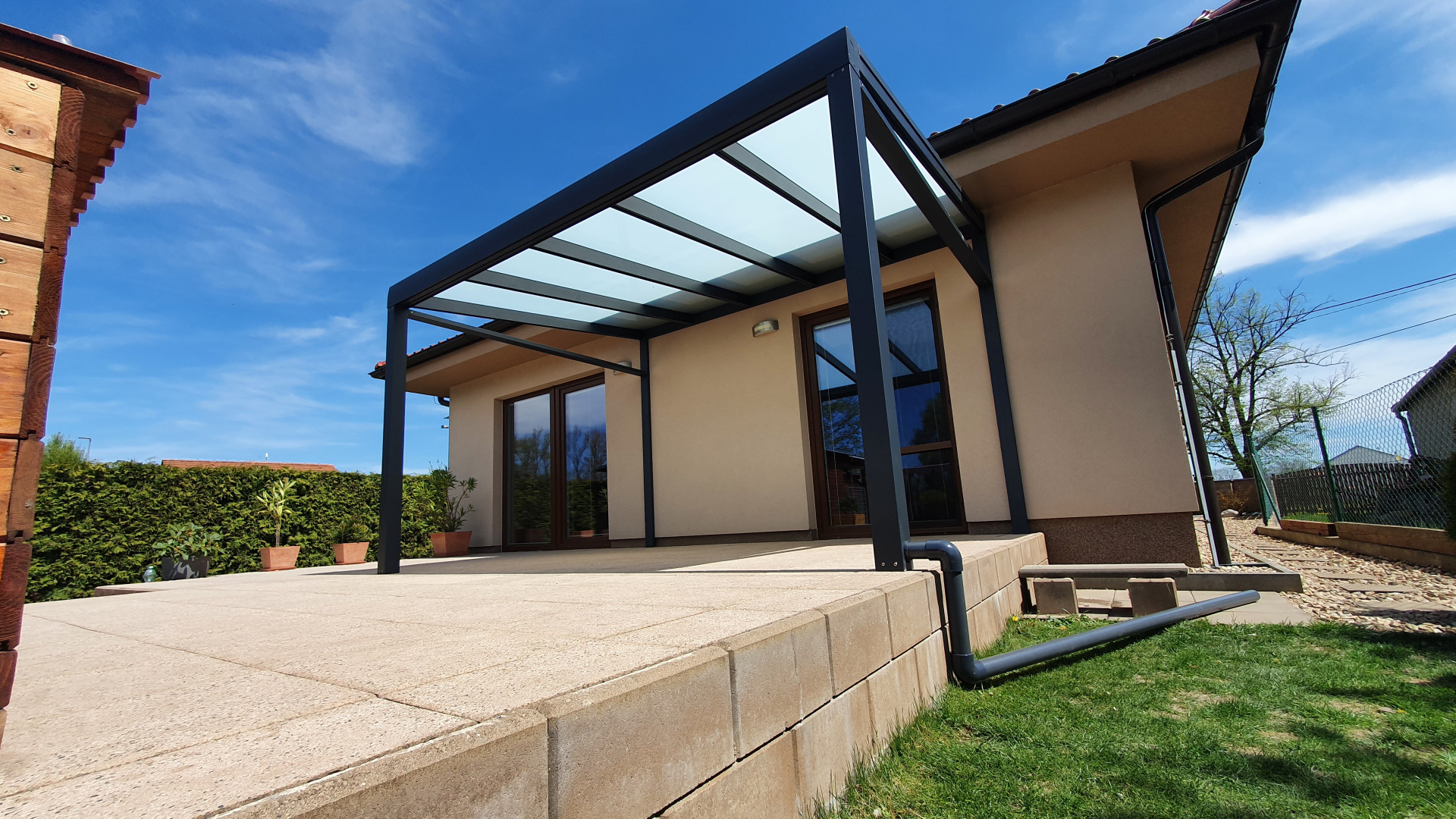Une pergola moderne en aluminium gris foncé avec un toit en verre, installée contre une maison individuelle sur une terrasse pavée.