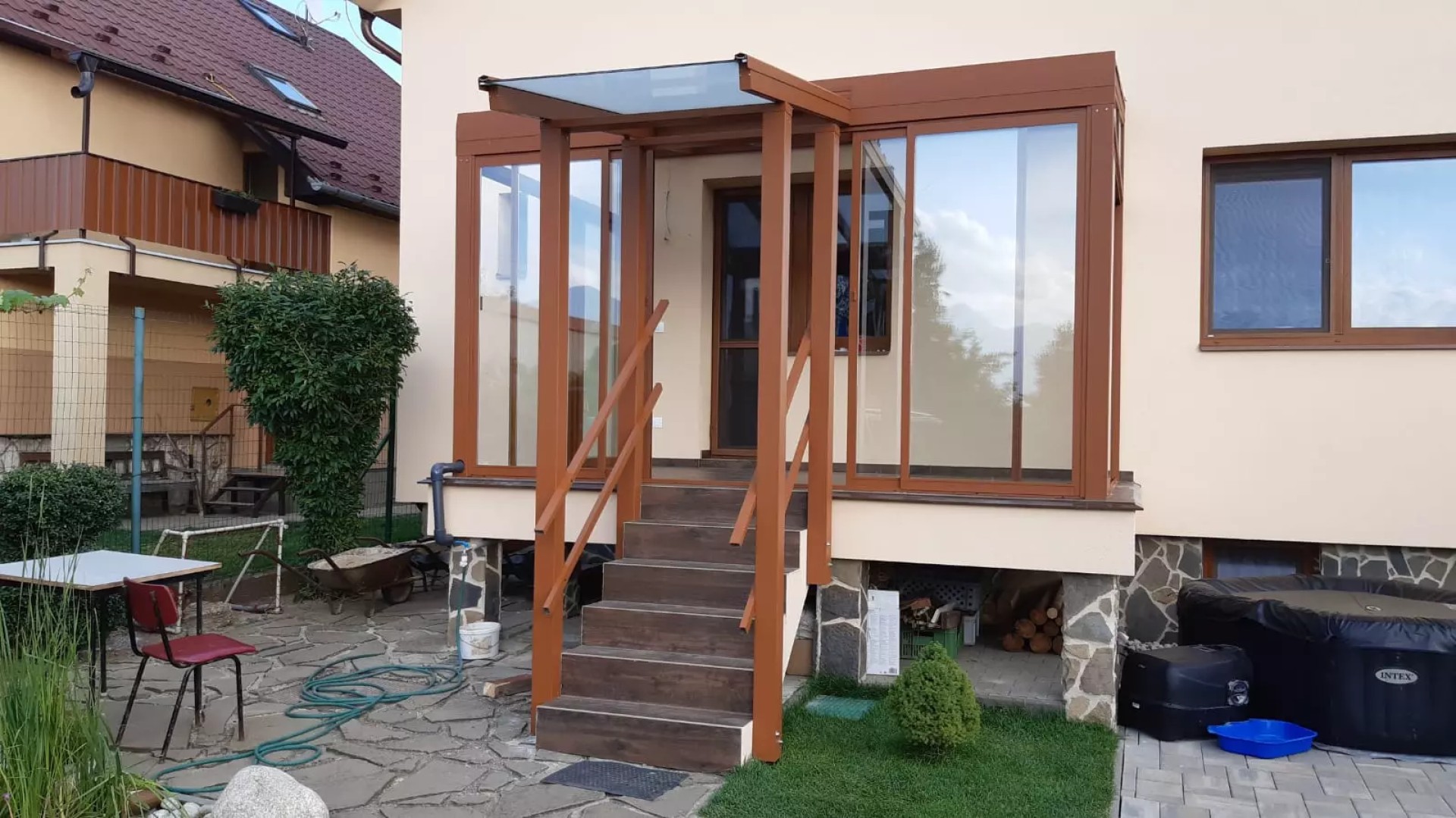 Pergola en aluminium vitrée de couleur marron, avec un auvent au-dessus des marches de l'entrée d'une maison individuelle.