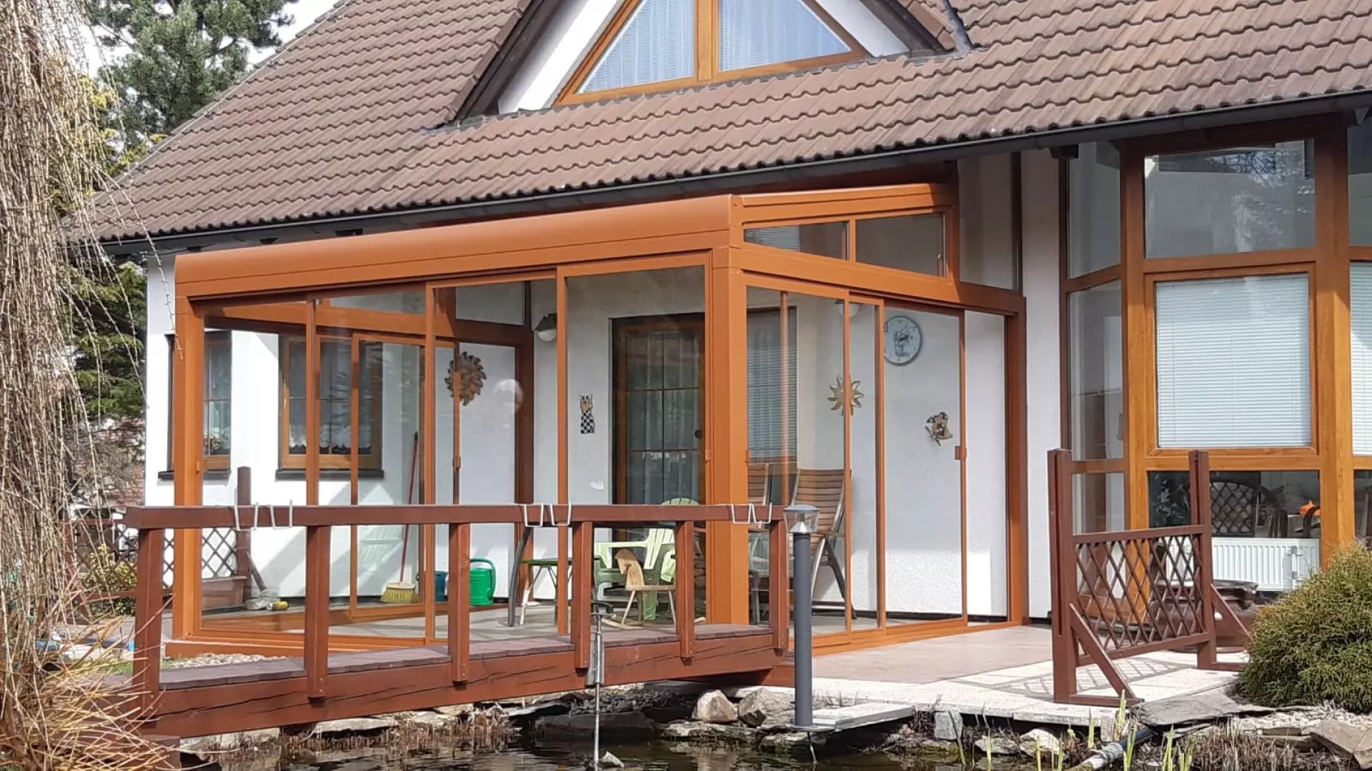 Pergola ROBUST en aluminium vitrée, de couleur marron, près d'une maison individuelle dotée d'une petite passerelle en bois