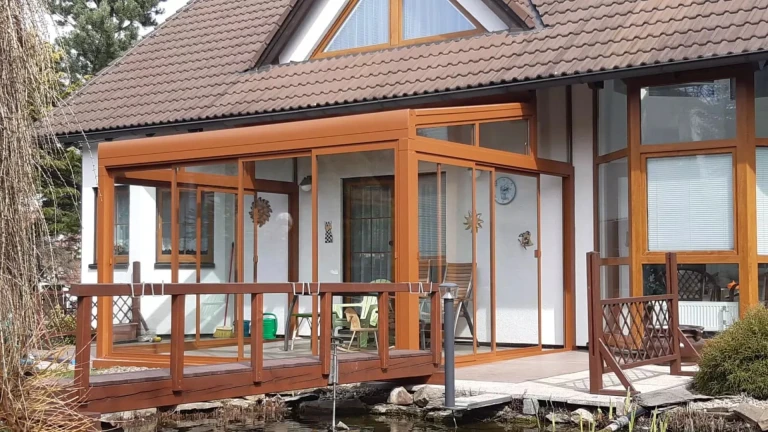 Pergola ROBUST en aluminium vitrée, de couleur marron, près d'une maison individuelle dotée d'une petite passerelle en bois