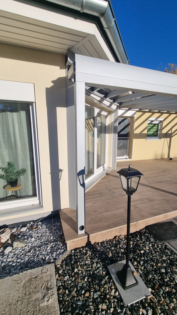 Détail de la fixation d'une pergola blanche en aluminium à la façade de la maison, sur une terrasse au décor boisé.