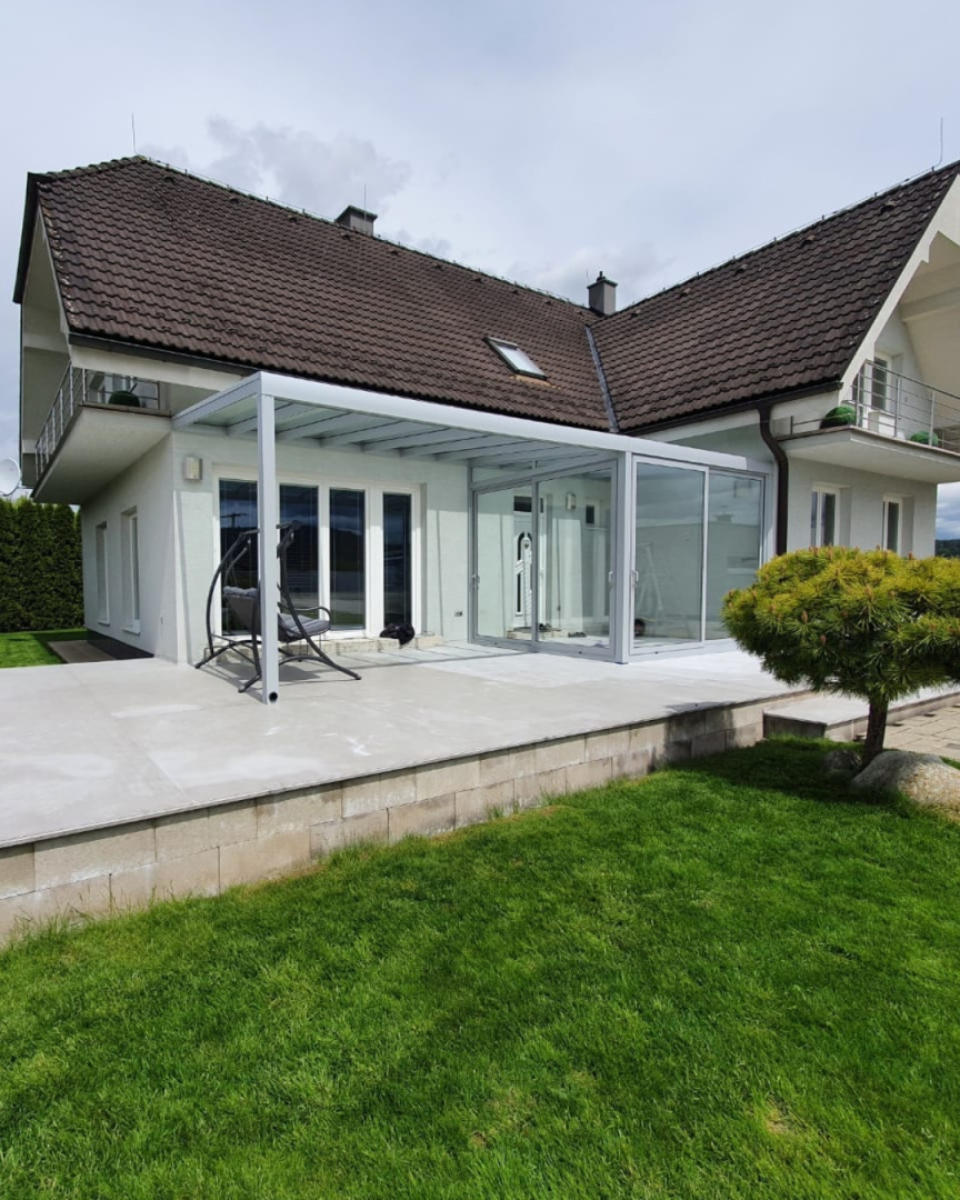 Une véranda moderne en aluminium, dotée de parois coulissantes vitrées, construite à côté d'une maison individuelle dotée d'une terrasse.
