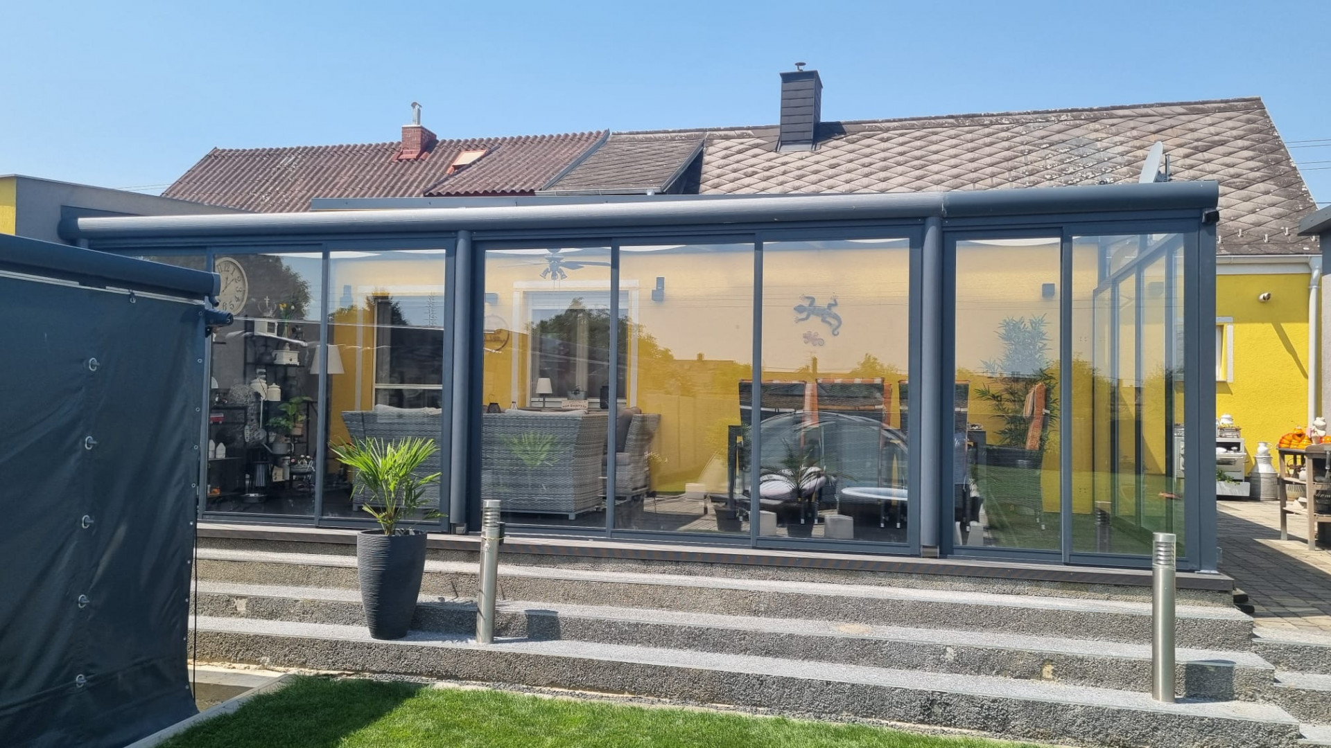 Une véranda moderne en aluminium et verre, de couleur anthracite, adossée à une maison individuelle jaune dotée d'un escalier.