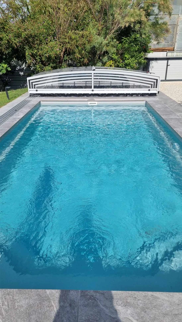 Vue sur la piscine en plein air, dont la petite verrière a été rabattue à l'extrémité de la piscine, dans le jardin