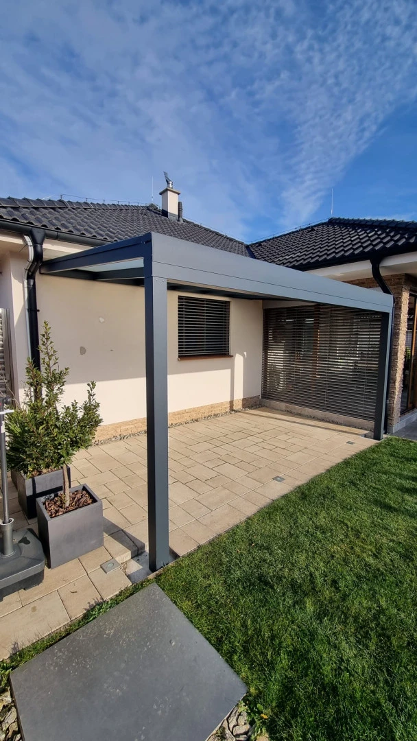 Une pergola moderne en aluminium anthracite, adossée à la maison, sur une terrasse pavée agrémentée d'une pelouse.