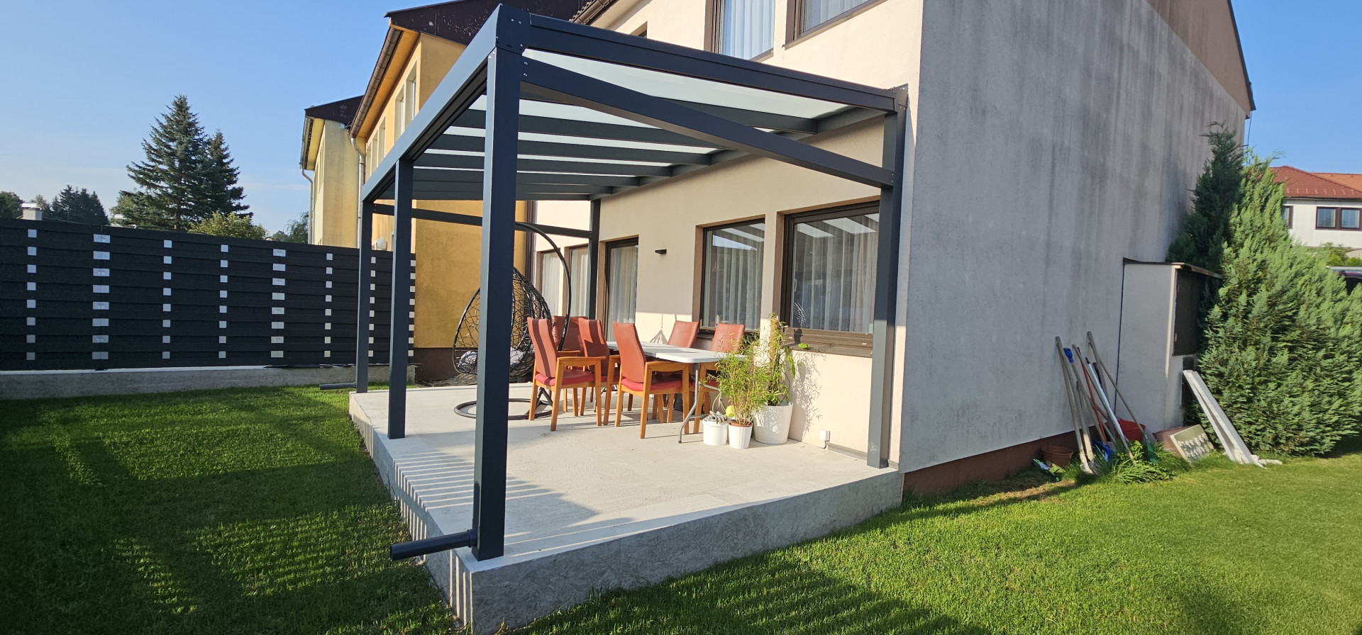 Une pergola moderne en aluminium avec un toit vitré, installée sur la terrasse en béton d'une maison individuelle, agrémentée de mobilier de jardin.