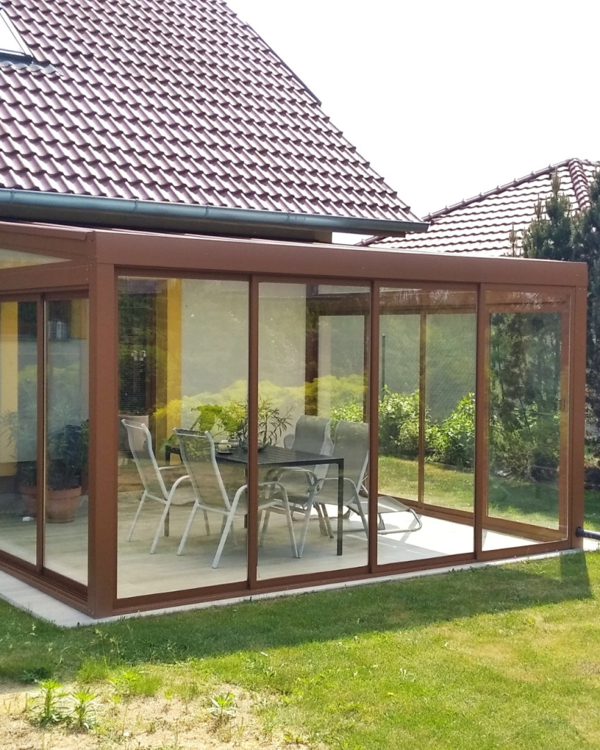 Une véranda vitrée en aluminium avec un cadre marron et du mobilier de jardin sur la terrasse attenante à la maison