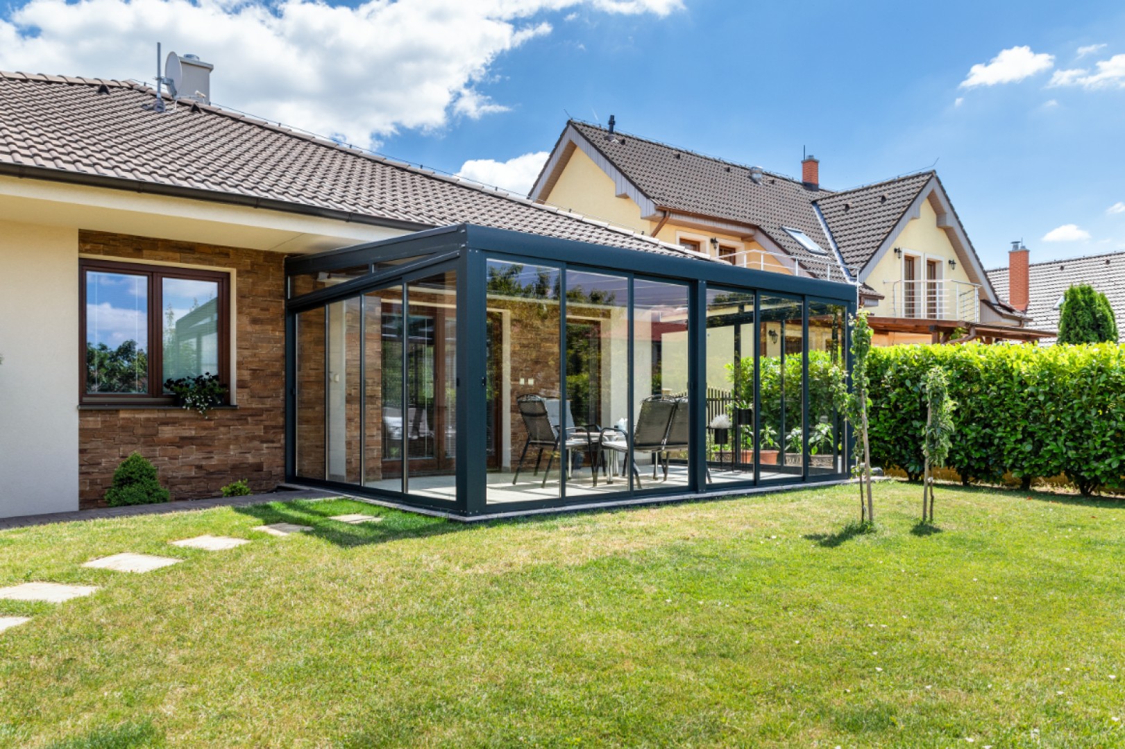 Une véranda moderne vitrée, dotée d'une structure sombre, attenante à une maison individuelle et équipée de mobilier de jardin.