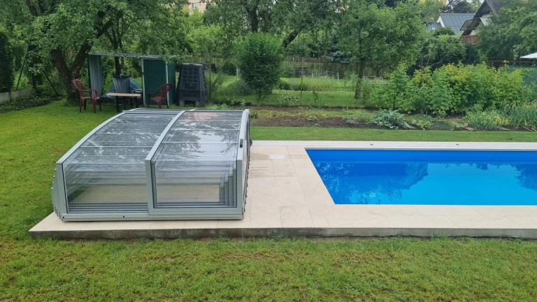 Vue sur la piscine bleue, dont la couverture grise et basse est relevée, dans le jardin.