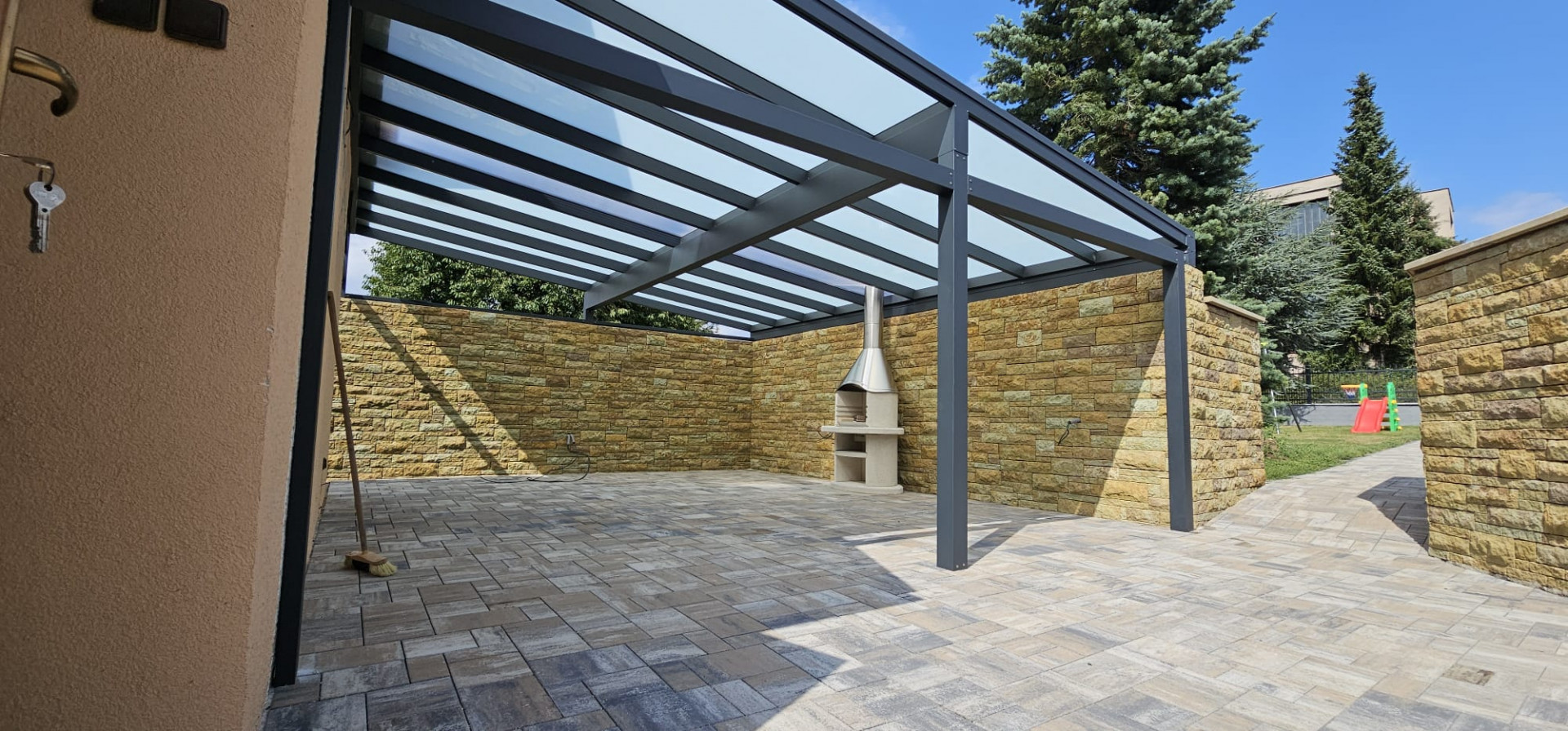 Une pergola moderne en aluminium pour la terrasse, avec une partie centrale vitrée, des murs revêtus de pierre et une cheminée d'extérieur.