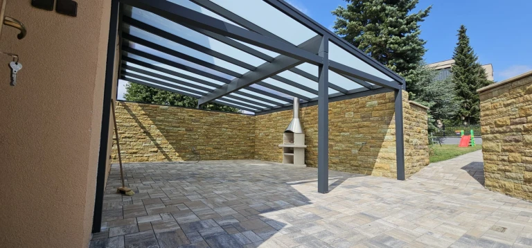 Une pergola moderne en aluminium pour la terrasse, avec une partie centrale vitrée, des murs revêtus de pierre et une cheminée d'extérieur.