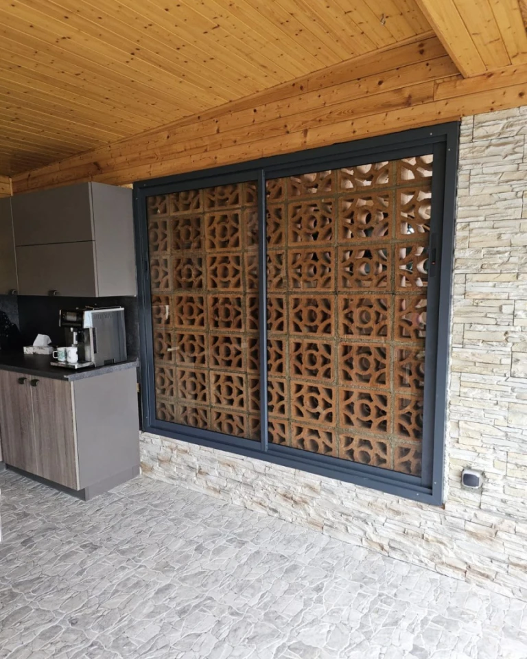 Une baie vitrée avec un cadre anthracite sur une terrasse dallée de pierre, offrant une vue sur un mur décoratif en briques.