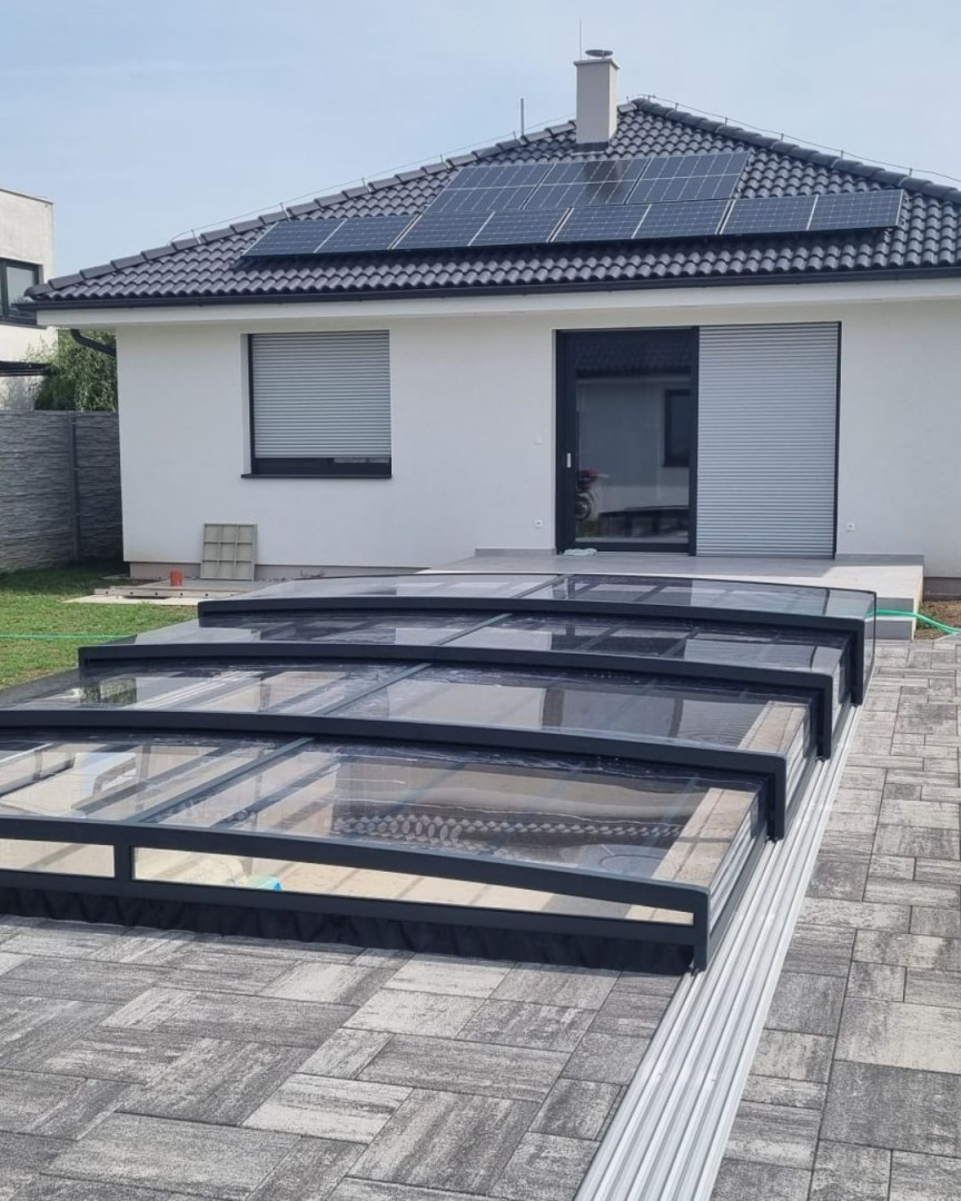 Une couverture de piscine moderne et basse, de couleur anthracite, devant une maison individuelle dotée d'un dallage et d'une installation photovoltaïque.
