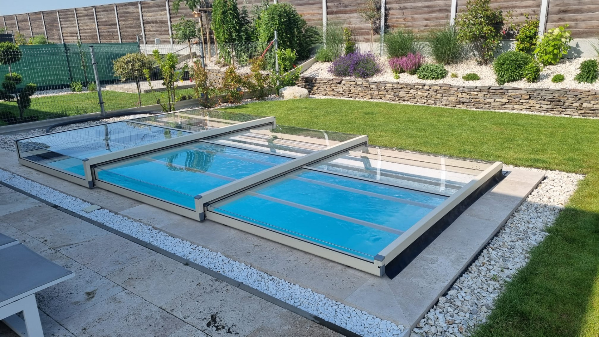 Une couverture télescopique moderne et basse pour piscine, dotée de segments vitrés, dans un jardin aménagé.