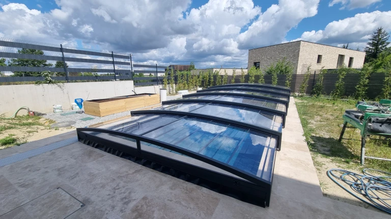 Une couverture de piscine moderne et basse, avec une structure noire et un revêtement transparent, dans le jardin d'une maison individuelle.