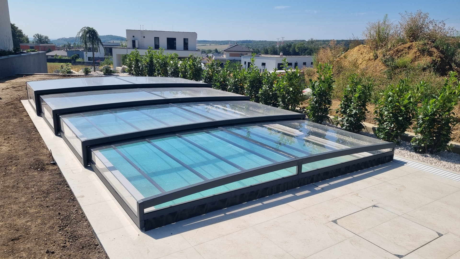 Une couverture de piscine télescopique moderne et basse, avec une structure anthracite et un revêtement transparent, installée sur la terrasse d'une maison individuelle.