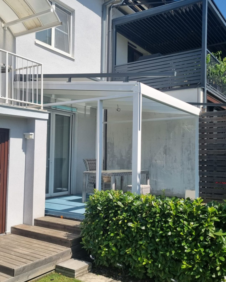 Une pergola moderne en aluminium blanc avec un toit en verre, près d'une maison individuelle équipée de mobilier de terrasse.