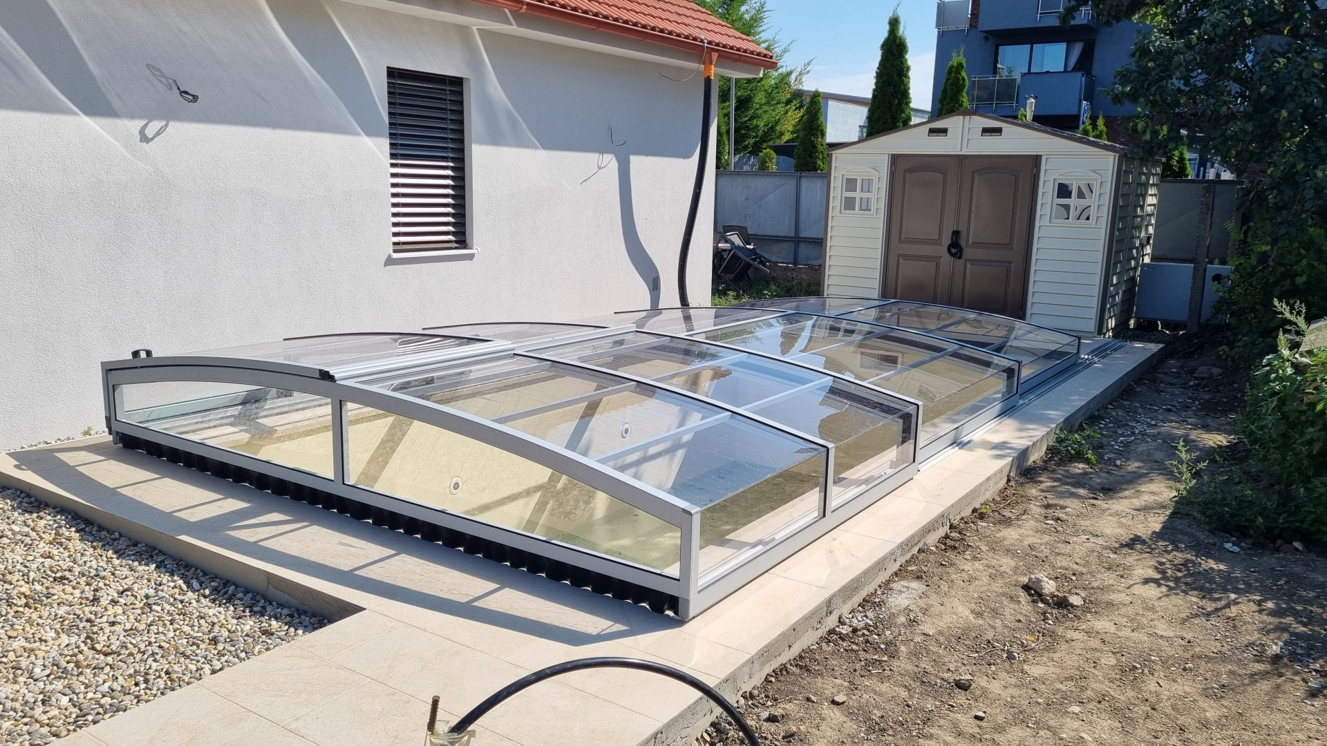 Une couverture de piscine basse à panneaux, avec une structure argentée et du polycarbonate transparent, près d'une maison individuelle à Senec.